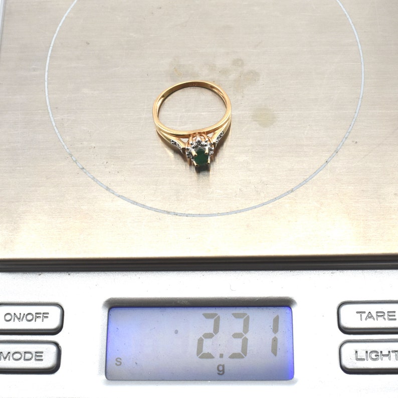 May include: A gold ring featuring a central oval-cut green gemstone surrounded by small clear stones, resting on a digital scale that displays a weight of 2.31 grams. The band is thin and polished, showcasing a classic design with a prong setting.