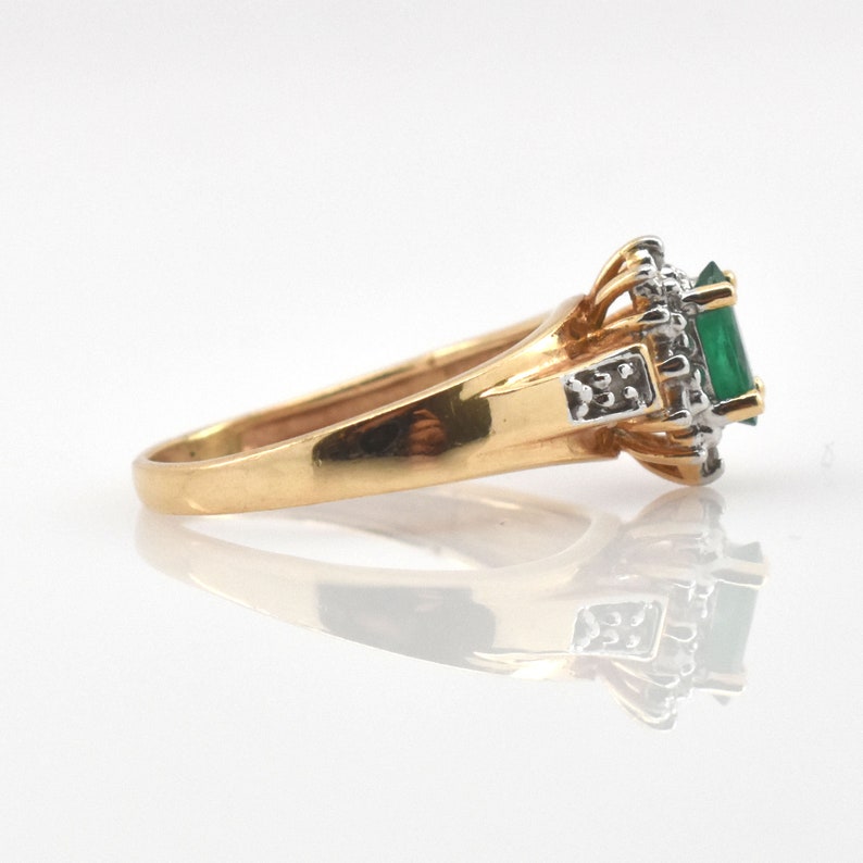 May include: A gold ring featuring a central emerald-cut green gemstone surrounded by a halo of small white diamonds. The setting is crafted from yellow gold with white gold accents near the stone, showcasing a polished band and side details.