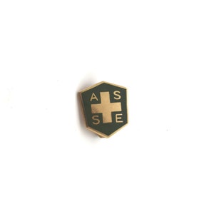 May include: A gold-colored pin with a green shield-shaped design. The shield has a white cross in the center and the letters "ASSE" around the cross.