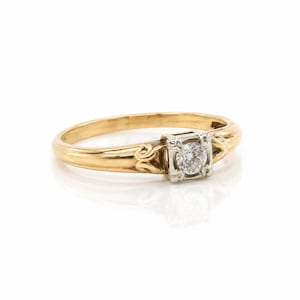 May include: A gold ring featuring a single round brilliant-cut diamond set in a square-shaped white metal head. The band is crafted from yellow gold and displays decorative scrollwork details on both sides of the central diamond setting.