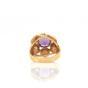 May include: A gold ring with a large, oval-cut purple gemstone. The ring has a detailed, textured band and an ornate setting with small accent stones. The ring is set against a white background, highlighting its intricate design and colour.