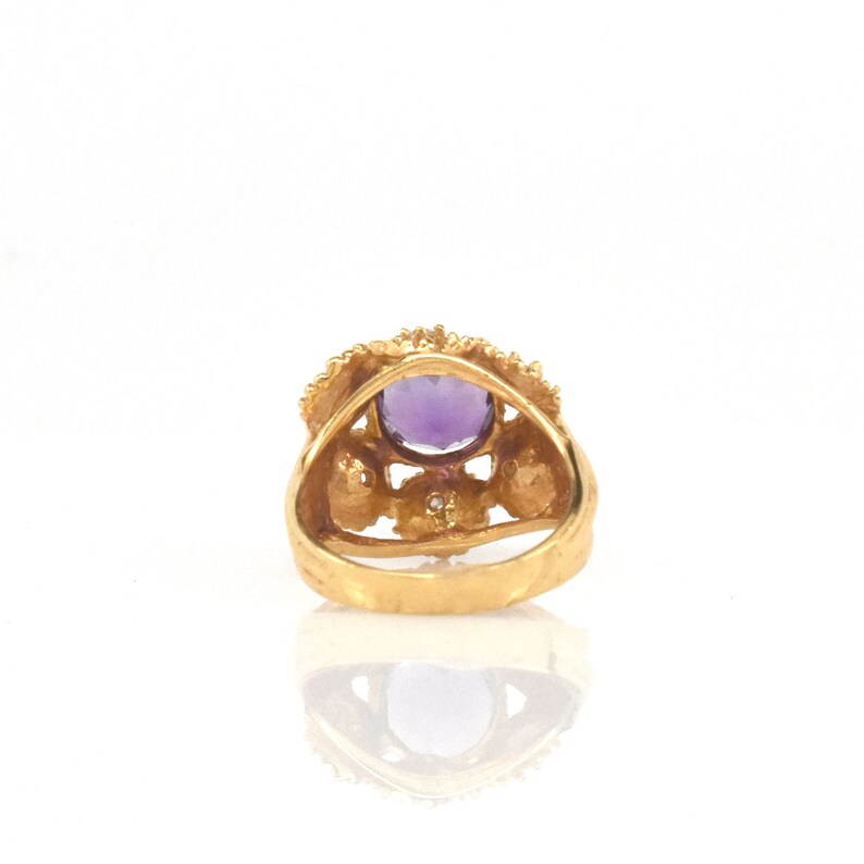 May include: A gold ring with a large, oval-cut purple gemstone. The ring has a detailed, textured band and an ornate setting with small accent stones. The ring is set against a white background, highlighting its intricate design and colour.