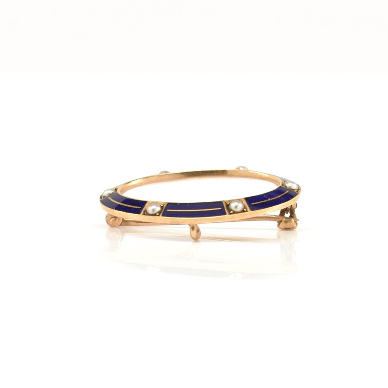 May include: A gold-toned brooch with a blue enamel design and pearl accents. The brooch is oval-shaped and has a hinged clasp.