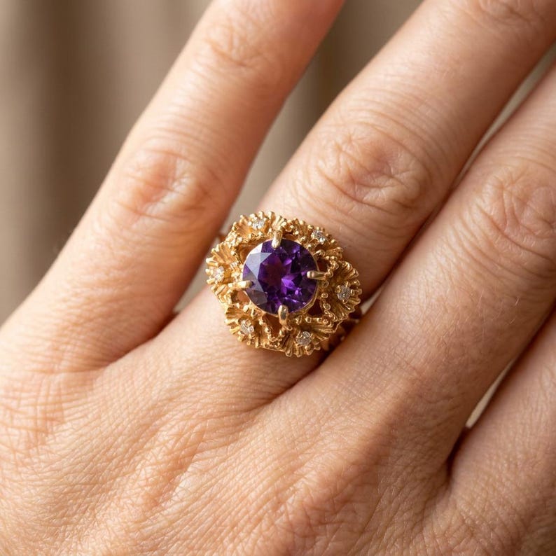 May include: A gold ring with a large, round, deep purple gemstone at the centre, encircled by smaller clear stones. The ring has an ornate, textured design with a floral pattern.