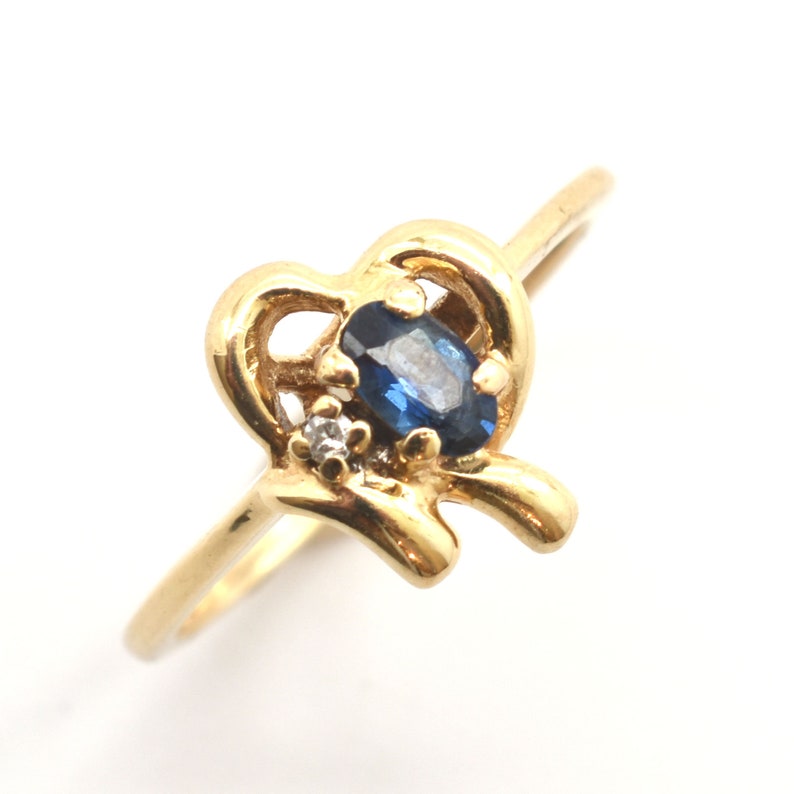 May include: A gold ring featuring a polished band that transitions into a stylized open heart shape. The setting holds a central oval-cut deep blue sapphire gemstone, accented by a single small, clear round-cut diamond positioned to the side.