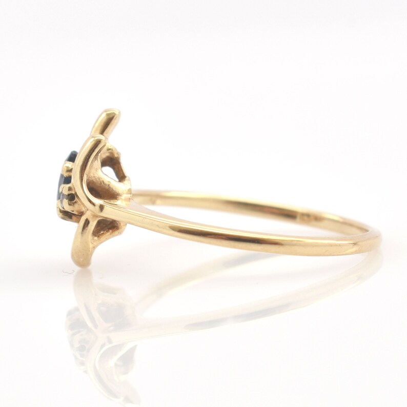 May include: A gold ring featuring a dark blue gemstone set in a prong setting. The band is smooth and polished, with a side profile view showing the elevated mounting of the stone and the curved metal structure that holds the gem securely in place.