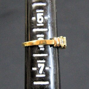 May include: A gold ring featuring a clear, round-cut gemstone set on a band. The ring is positioned on a black ring mandrel marked with white size indicators, showing the band rests between size 6 and 7 on the standard US ring sizing scale.