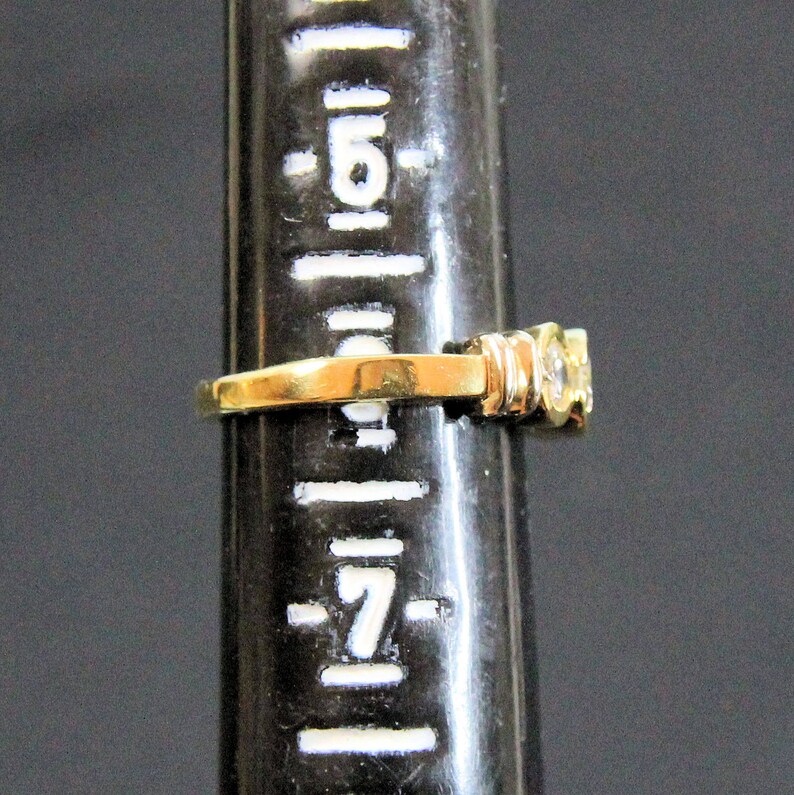 May include: A gold ring featuring a clear, round-cut gemstone set on a band. The ring is positioned on a black ring mandrel marked with white size indicators, showing the band rests between size 6 and 7 on the standard US ring sizing scale.