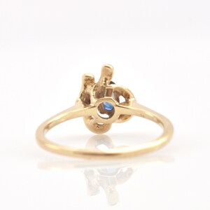 May include: A gold ring featuring a central oval-shaped blue gemstone set within an intricate, organic-shaped metal mounting. The band is smooth and circular, connecting to the textured, irregular metal design that holds the blue stone securely.
