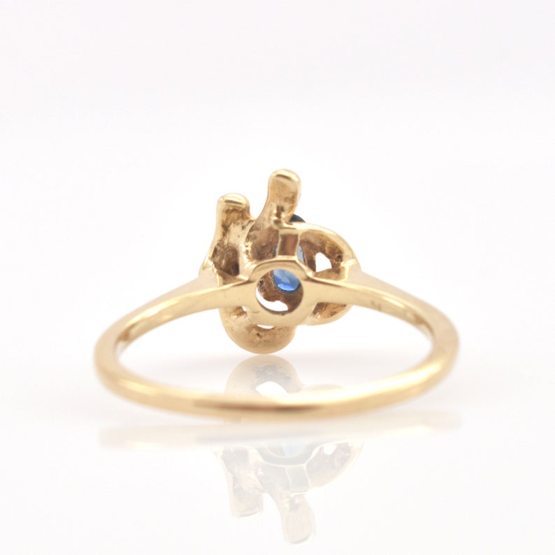 May include: A gold ring featuring a central oval-shaped blue gemstone set within an intricate, organic-shaped metal mounting. The band is smooth and circular, connecting to the textured, irregular metal design that holds the blue stone securely.