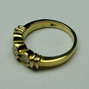 May include: A yellow gold ring featuring three round, bezel-set clear stones arranged in a row. The interior of the band is clearly stamped with the text 18K, indicating the gold purity. The metal has a polished, reflective finish throughout.