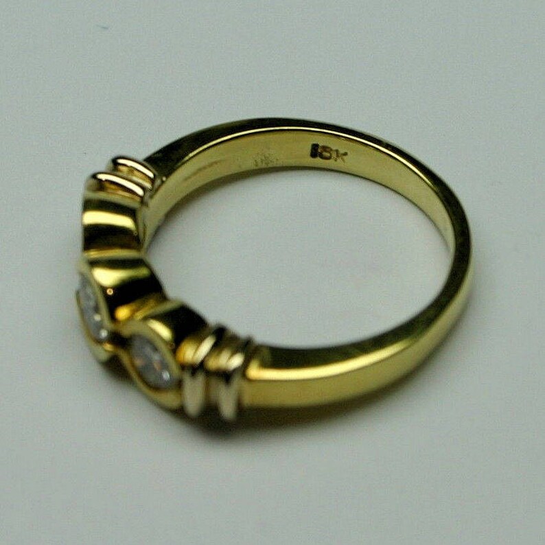 May include: A yellow gold ring featuring three round, bezel-set clear stones arranged in a row. The interior of the band is clearly stamped with the text 18K, indicating the gold purity. The metal has a polished, reflective finish throughout.