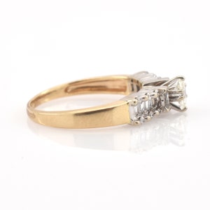 May include: A gold ring with a large, clear diamond center stone and smaller, rectangular diamonds on either side. The ring has a classic design with a polished gold band and a white metal setting for the diamonds. The ring is set against a white background.