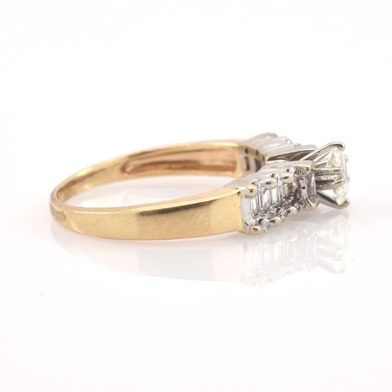 May include: A gold ring with a large, clear diamond center stone and smaller, rectangular diamonds on either side. The ring has a classic design with a polished gold band and a white metal setting for the diamonds. The ring is set against a white background.