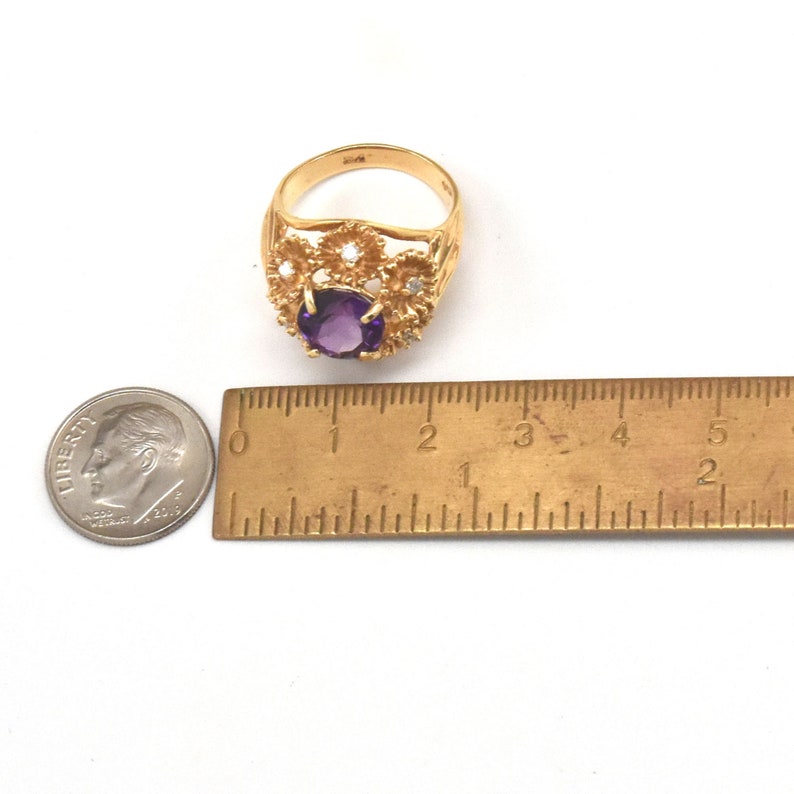 May include: A gold ring with a large, round, purple gemstone at the centre, encircled by smaller white stones and floral filigree. The ring is shown with a ruler and a coin for size reference.
