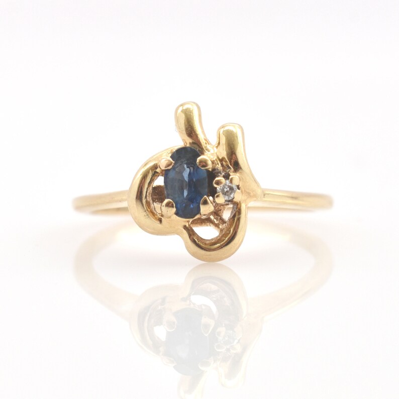 May include: A gold ring featuring an oval-cut dark blue sapphire and a small round-cut clear diamond set within an asymmetrical, swirling metal band. The polished yellow gold setting curves around the stones, creating a fluid, open design.