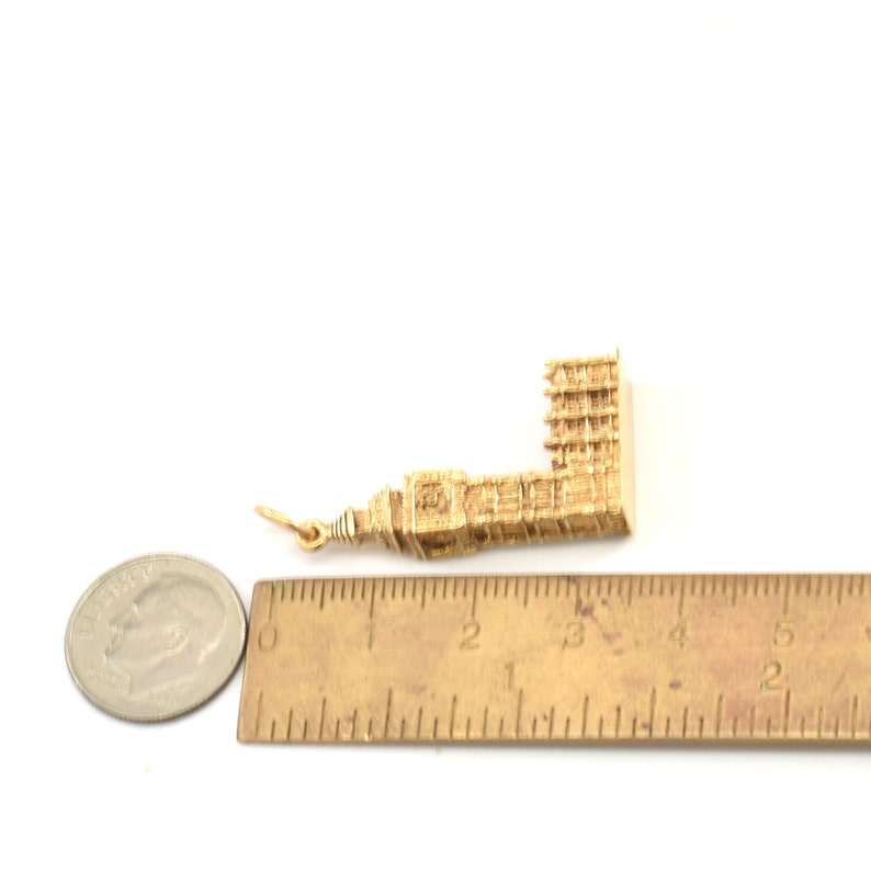 May include: A gold-colored charm in the shape of a building, measuring approximately 3 inches long. The charm is next to a ruler for size comparison.