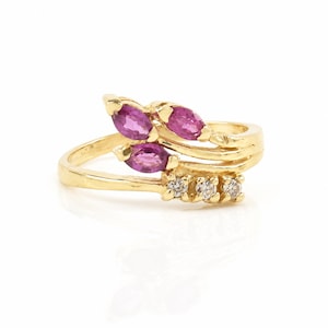 May include: A gold ring featuring two marquise-cut pink gemstones arranged in a bypass style, complemented by a row of three small, clear round-cut stones set along the lower band, creating a layered and asymmetrical design with a polished finish.