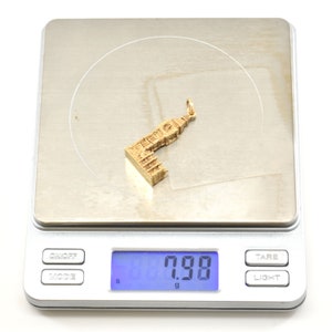 May include: A gold-colored charm in the shape of a building, weighing 7.98 grams, on a digital scale.