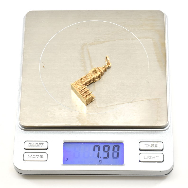 May include: A gold-colored charm in the shape of a building, weighing 7.98 grams, on a digital scale.