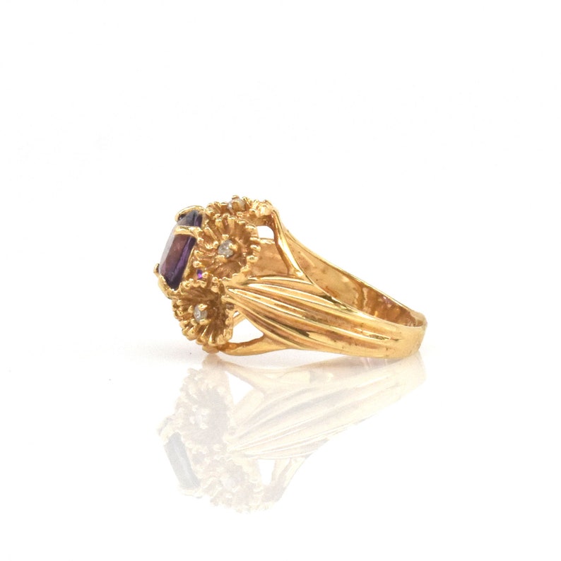May include: A gold ring with a large, oval-cut purple gemstone. The ring has a floral design with small, clear accent stones and detailed goldwork. The band is smooth and polished. The ring's design is intricate and eye-catching.
