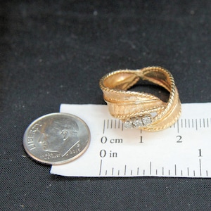 May include: Gold-toned ring with a textured, ribbon-like design. The ring features three small, clear stones and a rope-like border. A U.S. dime is next to the ring for size comparison.