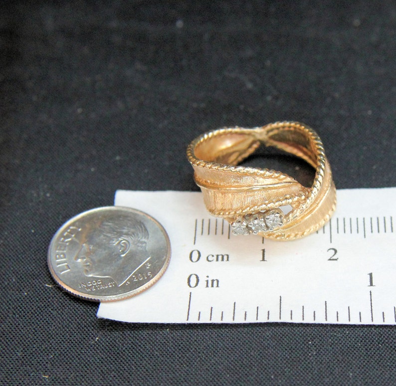 May include: Gold-toned ring with a textured, ribbon-like design. The ring features three small, clear stones and a rope-like border. A U.S. dime is next to the ring for size comparison.