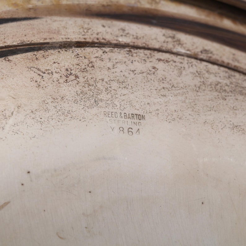 May include: Close-up of a silver serving dish with the text "REED & BARTON STERLING V 864" stamped on the surface. The dish has a textured, aged appearance with dark brown accents.