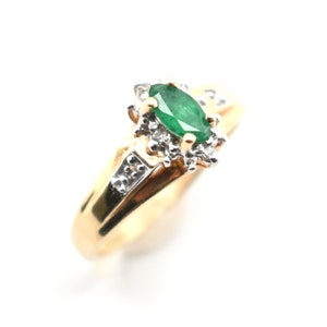 May include: A gold ring featuring a central marquise-cut green emerald stone surrounded by a cluster of small clear white stones. The band is made of polished yellow gold and includes additional small white stones set along the shoulders.