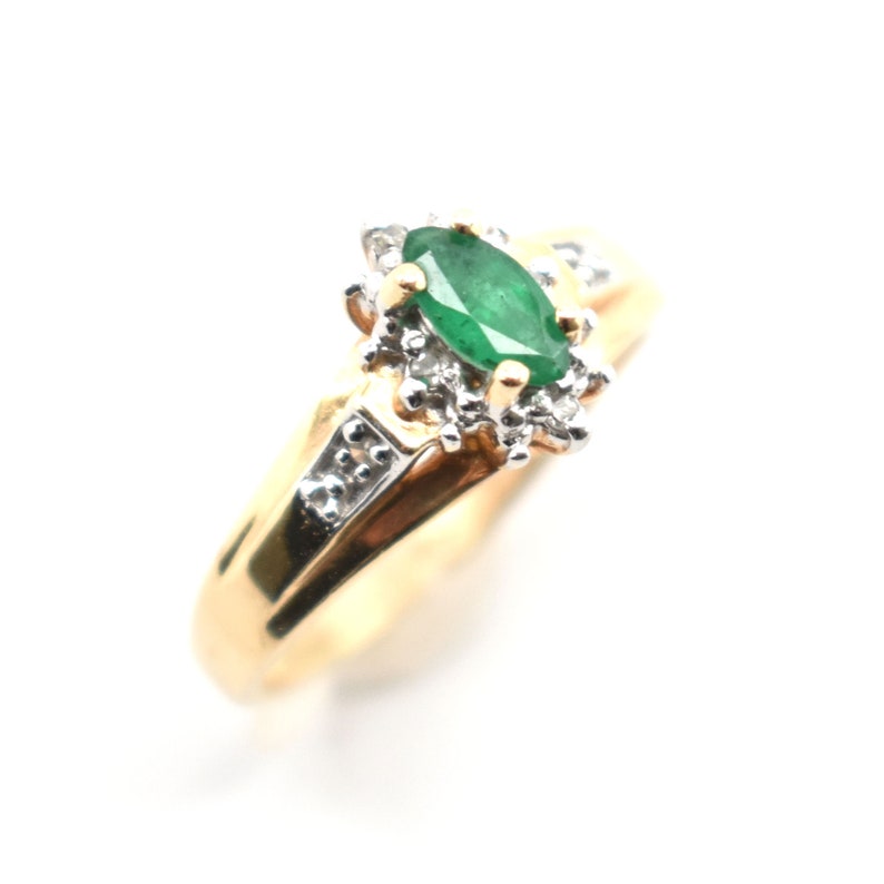 May include: A gold ring featuring a central marquise-cut green emerald stone surrounded by a cluster of small clear white stones. The band is made of polished yellow gold and includes additional small white stones set along the shoulders.