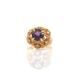 May include: A gold ring with a large, round, deep purple gemstone at its centre, encircled by smaller diamonds and a textured gold setting. The ring's design incorporates floral elements, giving it a vintage or antique appearance.