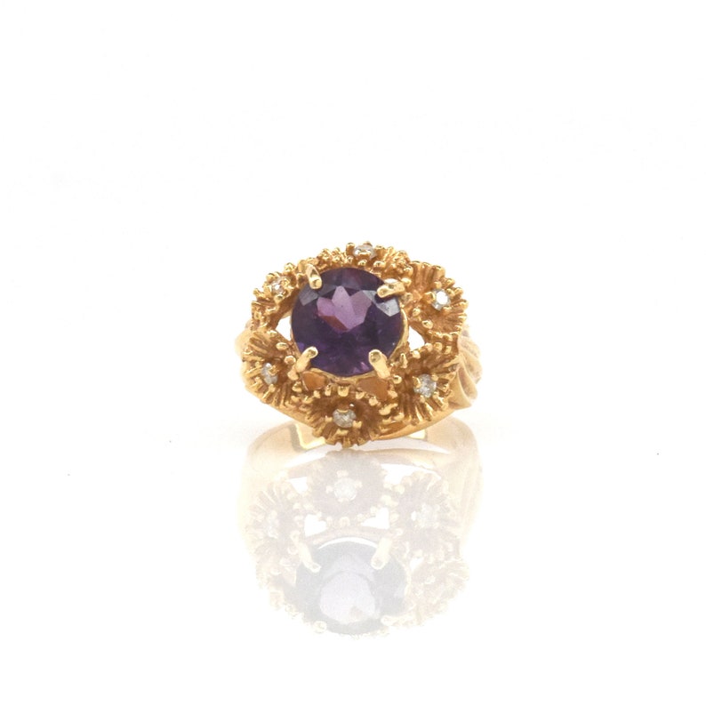 May include: A gold ring with a large, round, deep purple gemstone at its centre, encircled by smaller diamonds and a textured gold setting. The ring's design incorporates floral elements, giving it a vintage or antique appearance.