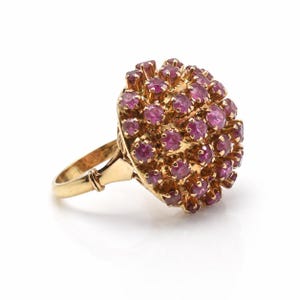 May include: A gold ring featuring a cluster of small, round, pink gemstones. The ring's band and setting are a warm gold color, and the gemstones are closely set together, forming a dome shape. The ring is set against a white background.