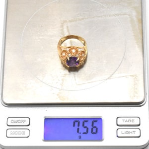 May include: A gold ring featuring a central oval, deep purple gemstone. The ring is adorned with smaller, clear stones and floral patterns. The ring is on a digital scale, displaying a weight of 7.56 grams.