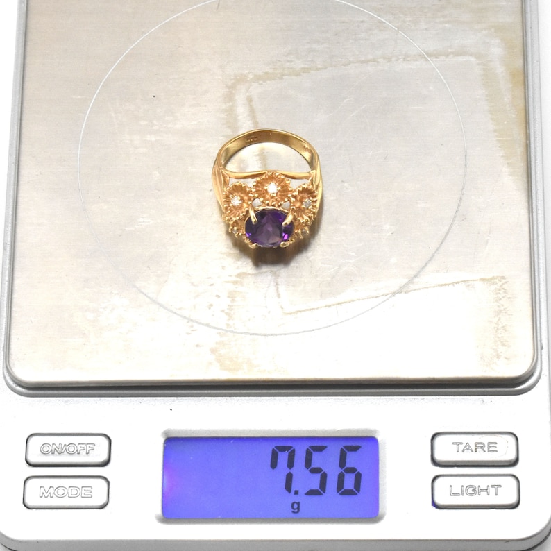 May include: A gold ring featuring a central oval, deep purple gemstone. The ring is adorned with smaller, clear stones and floral patterns. The ring is on a digital scale, displaying a weight of 7.56 grams.