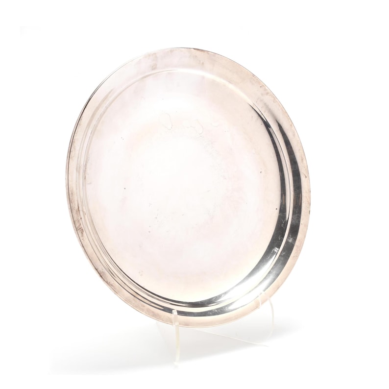 May include: A silver-colored, oval serving tray with a raised rim. The tray is displayed on a clear stand against a white background. The reflective surface of the tray catches the light, highlighting its smooth texture.