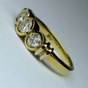 May include: A gold ring featuring three round, clear, faceted stones set in a row. The band is smooth and polished, with a distinct metal setting that encircles each stone individually, creating a continuous, connected design along the top.