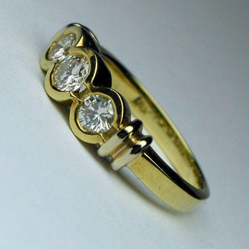 May include: A gold ring featuring three round, clear, faceted stones set in a row. The band is smooth and polished, with a distinct metal setting that encircles each stone individually, creating a continuous, connected design along the top.