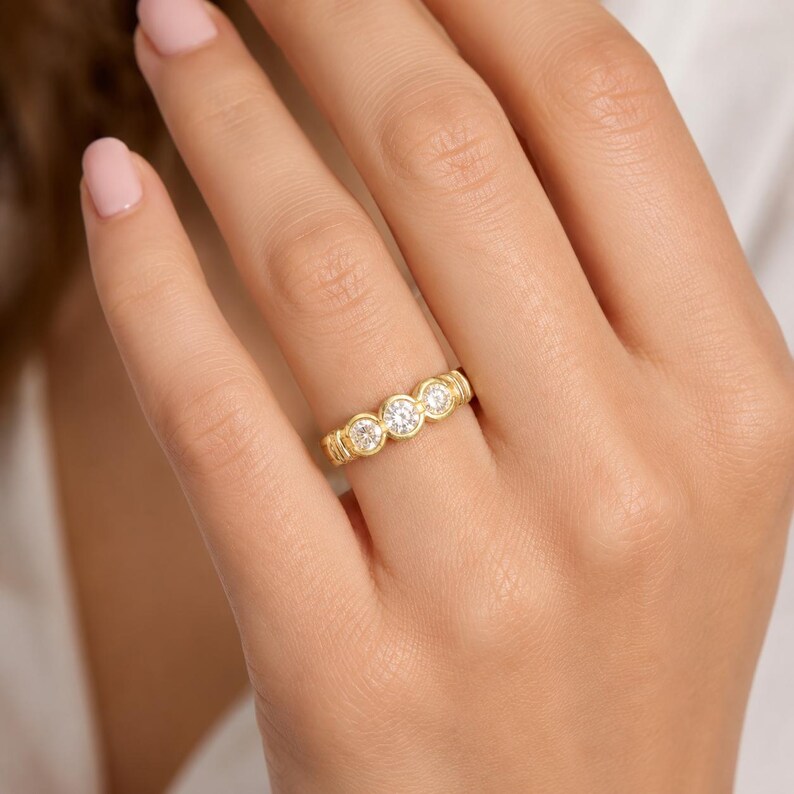 May include: A gold band ring featuring three round, clear, faceted stones set in individual circular bezels. The metal has a textured, ribbed finish along the band, and the stones are arranged horizontally across the center of the ring.