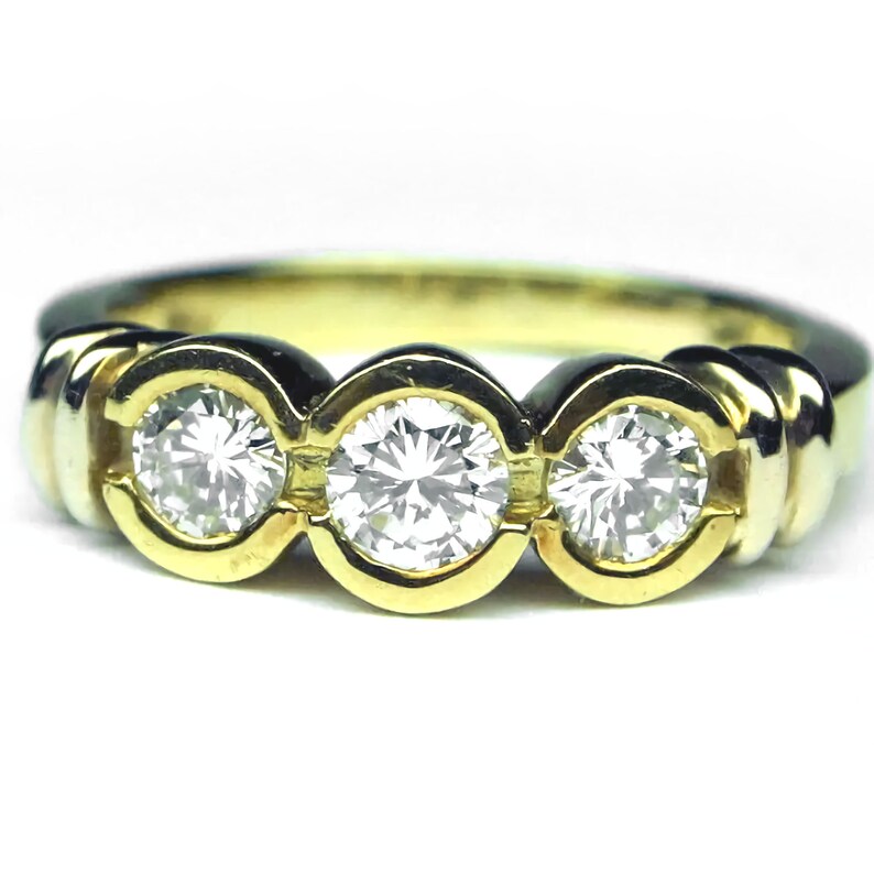 May include: A yellow gold ring featuring three round brilliant cut clear stones set in individual circular bezels. The band has a smooth, polished finish and a continuous design that highlights the three stones arranged horizontally across the top.