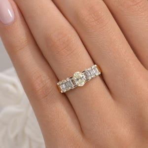 May include: A gold ring featuring an oval-cut diamond center stone, flanked by baguette-cut diamonds. The ring is set in a yellow gold band, showcasing a classic and elegant design. The diamonds sparkle, reflecting light.