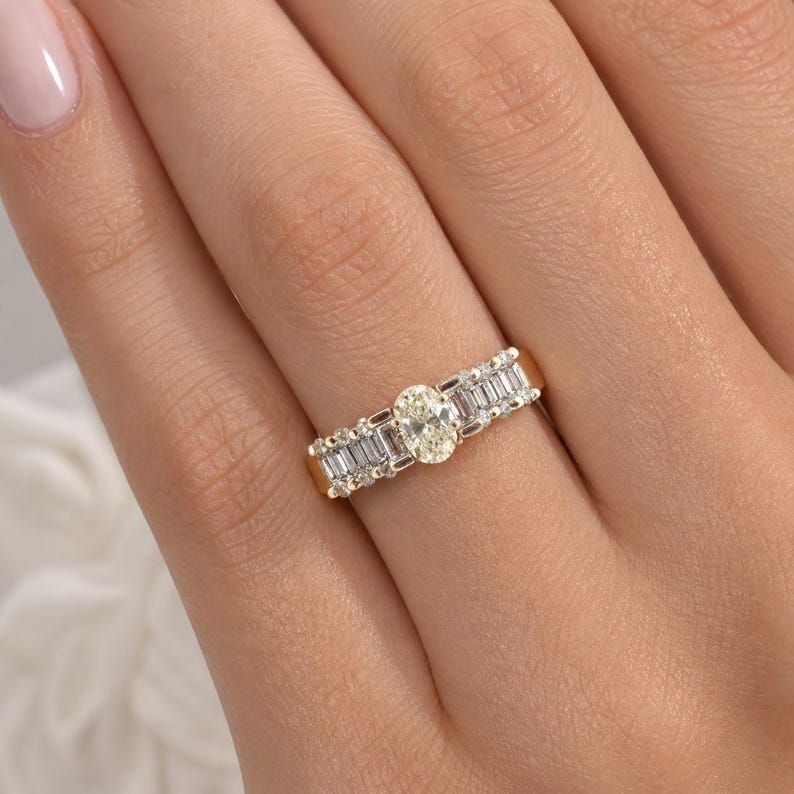 May include: A gold ring featuring an oval-cut diamond center stone, flanked by baguette-cut diamonds. The ring is set in a yellow gold band, showcasing a classic and elegant design. The diamonds sparkle, reflecting light.