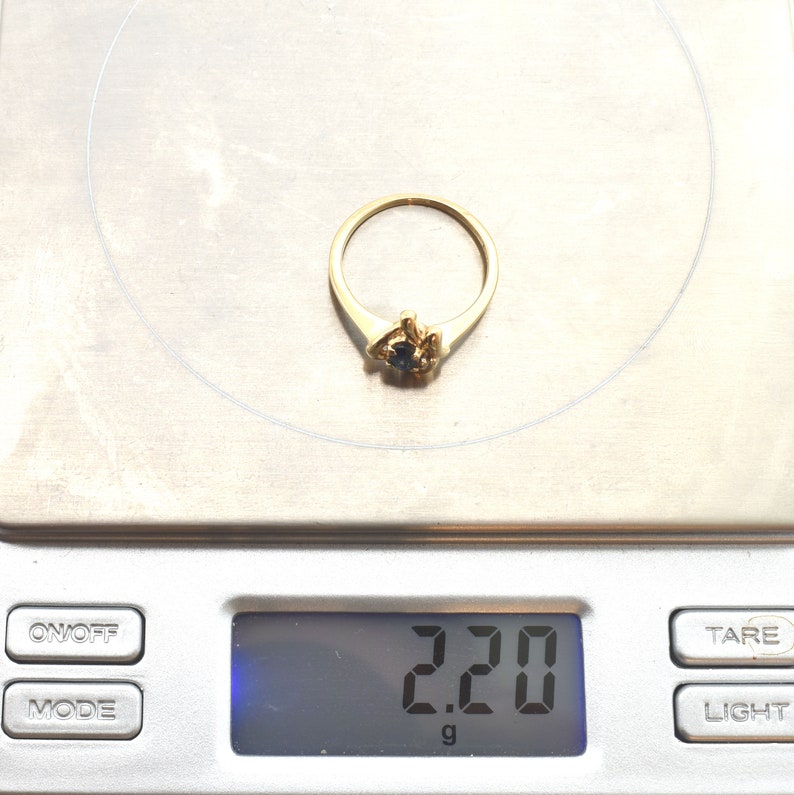 May include: A gold ring with a small dark blue gemstone set in a decorative mounting, resting on a digital scale that displays a weight of 2.20 grams. The ring features a thin, polished band and a central setting holding the round blue stone.