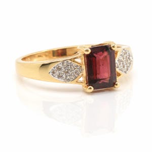 May include: A gold ring with a rectangular, deep red gemstone. The ring has diamond accents on either side of the main stone. The band is polished gold, and the ring is set against a white background.