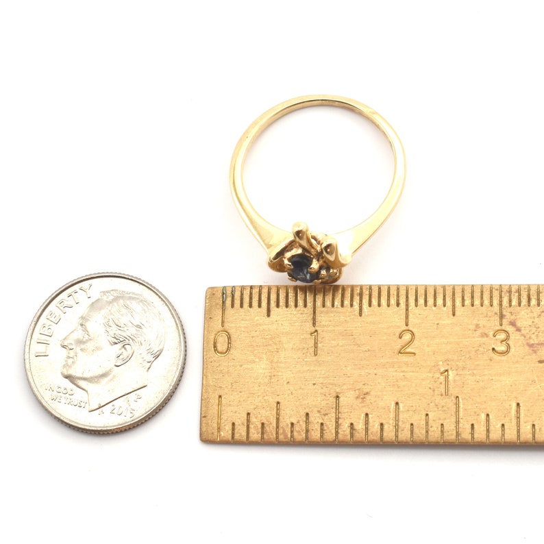 May include: A gold ring featuring a small, dark blue gemstone set in a decorative prong setting. The ring is positioned next to a metal ruler and a United States dime, showing the ring band has a diameter of approximately 0.75 inches.