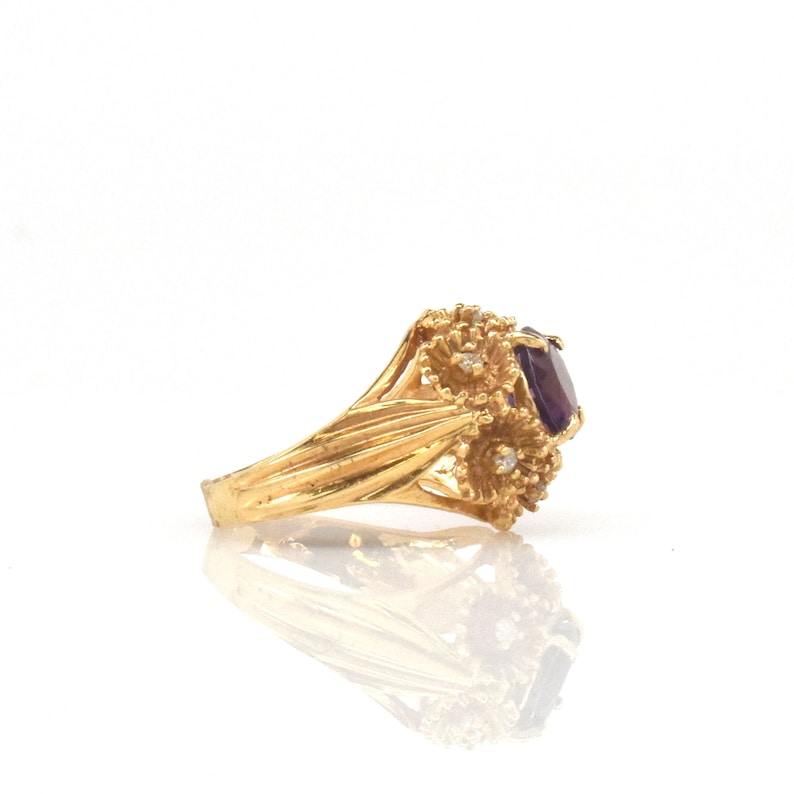 May include: A gold ring with a large, oval-cut purple gemstone. The ring has a floral design with small accent stones. The band is textured and set against a white background.