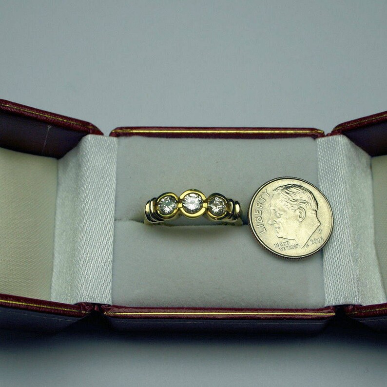 May include: A gold ring featuring three round, clear, faceted stones set in a horizontal row. The ring is positioned inside a red velvet-lined box next to a silver-colored United States dime for scale, which measures approximately 0.7 inches in diameter.