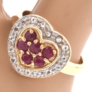 May include: A gold heart-shaped ring with a cluster of red rubies in the center and a halo of clear diamonds around the rubies.