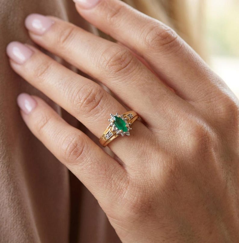 May include: A gold ring featuring a central marquise-cut green gemstone surrounded by a halo of small white stones. The band is crafted from yellow gold and includes additional small white stones set along the shoulders of the ring setting.