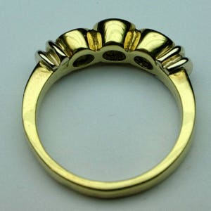 May include: A gold ring featuring a polished band and a setting that holds three circular stones. The metal has a smooth, reflective finish, and the design includes visible prongs securing each of the three stones along the top of the band.